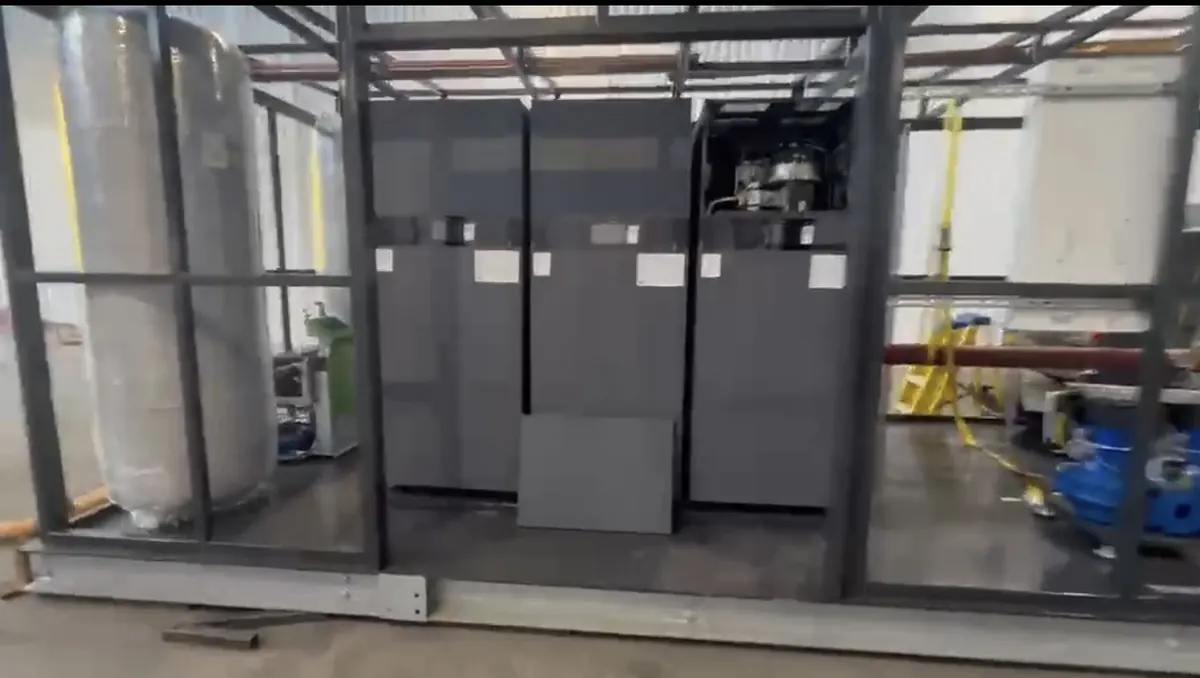 Front view of three tall grey floor-standing boiler cabinets inside a steel frame, with a lagged vessel on the left and pump sets and red-lagged pipework on the right
