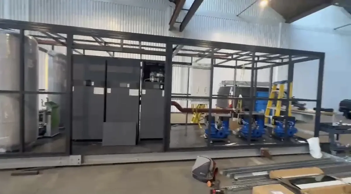 Wide view of a prefabricated steel-frame plant room in a workshop, with three grey floor-standing boiler cabinets, lagged vessels on the left and three blue pumps on the right